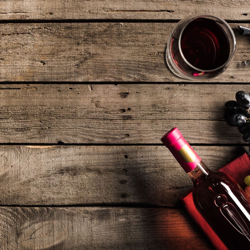 tablou canvas cu o sticla si un pahar de vin rosu, alaturi de struguri albi si negri, pe o masa de lemn, intr-un peisaj traditional