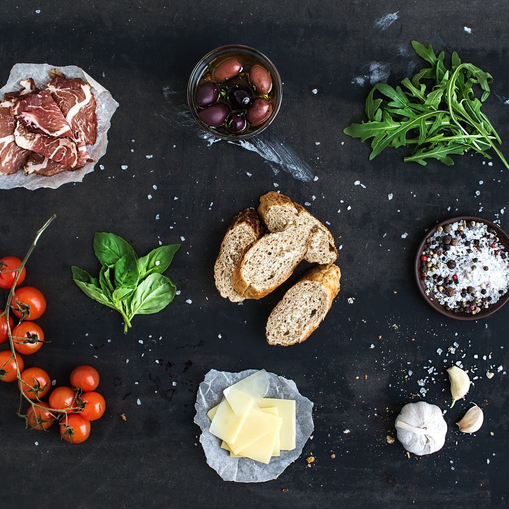 tablou canvas cu bruschette, rosii cherry, verdeturi, condimente si prosciutto pe o masa neagra