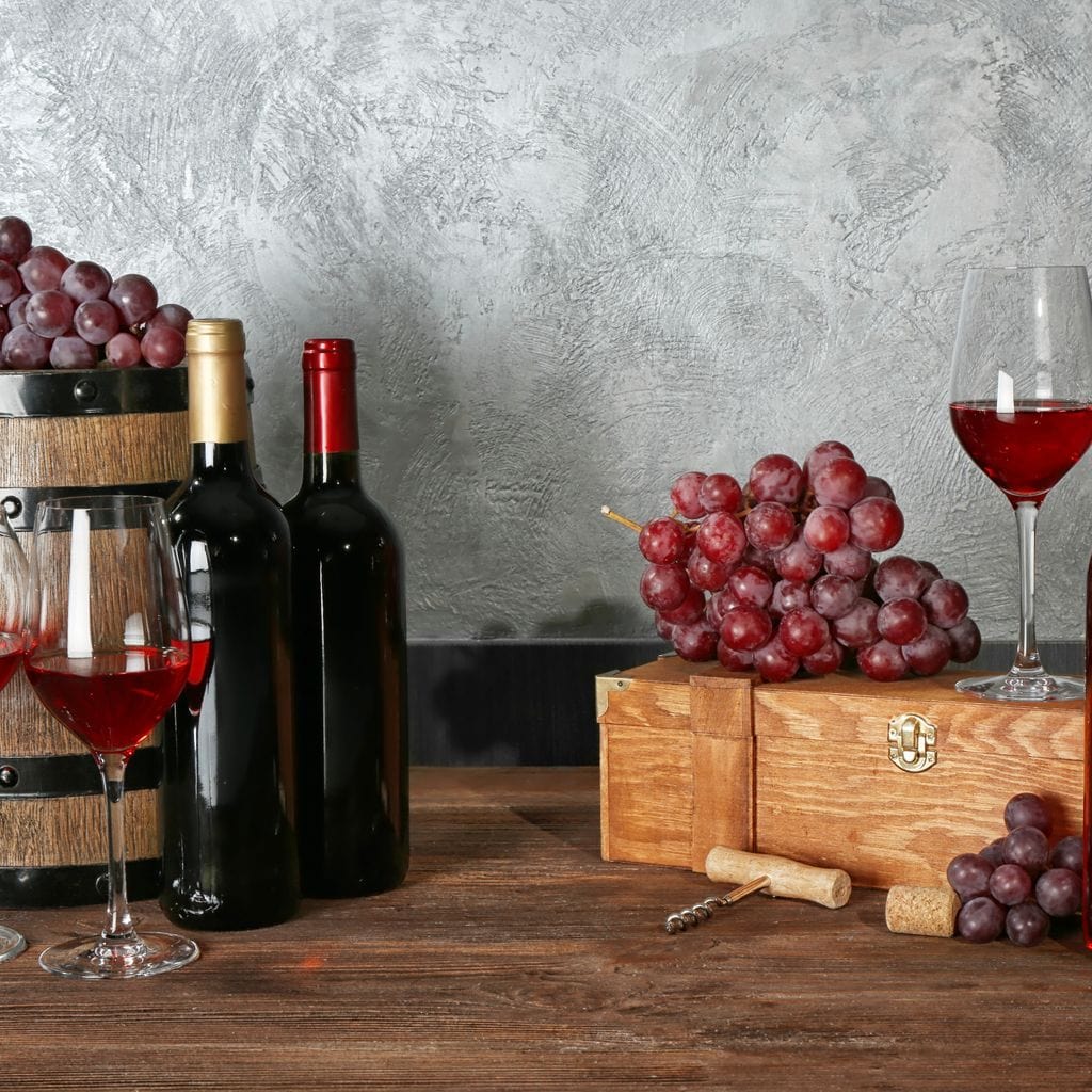 tablou canvas cu trei sticle de vin si pahare cu vin rosu pe o masa de lemn in stil rustic, alaturi de ciorchini de struguri
