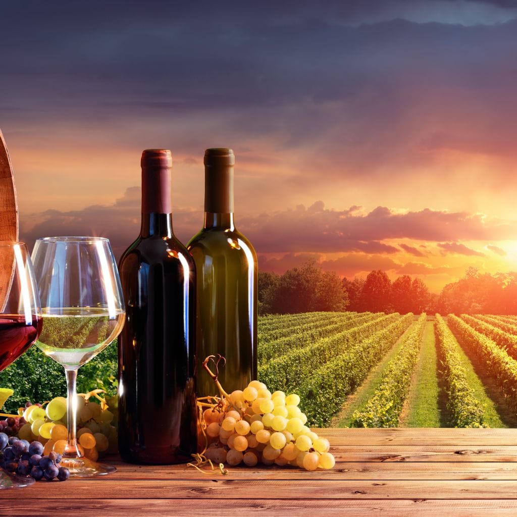 tablou canvas cu un butoi de lemn, doua sticle si doua pahare de vin, alaturi de struguri albi si rosii, pe fundal rustic la apus de soare