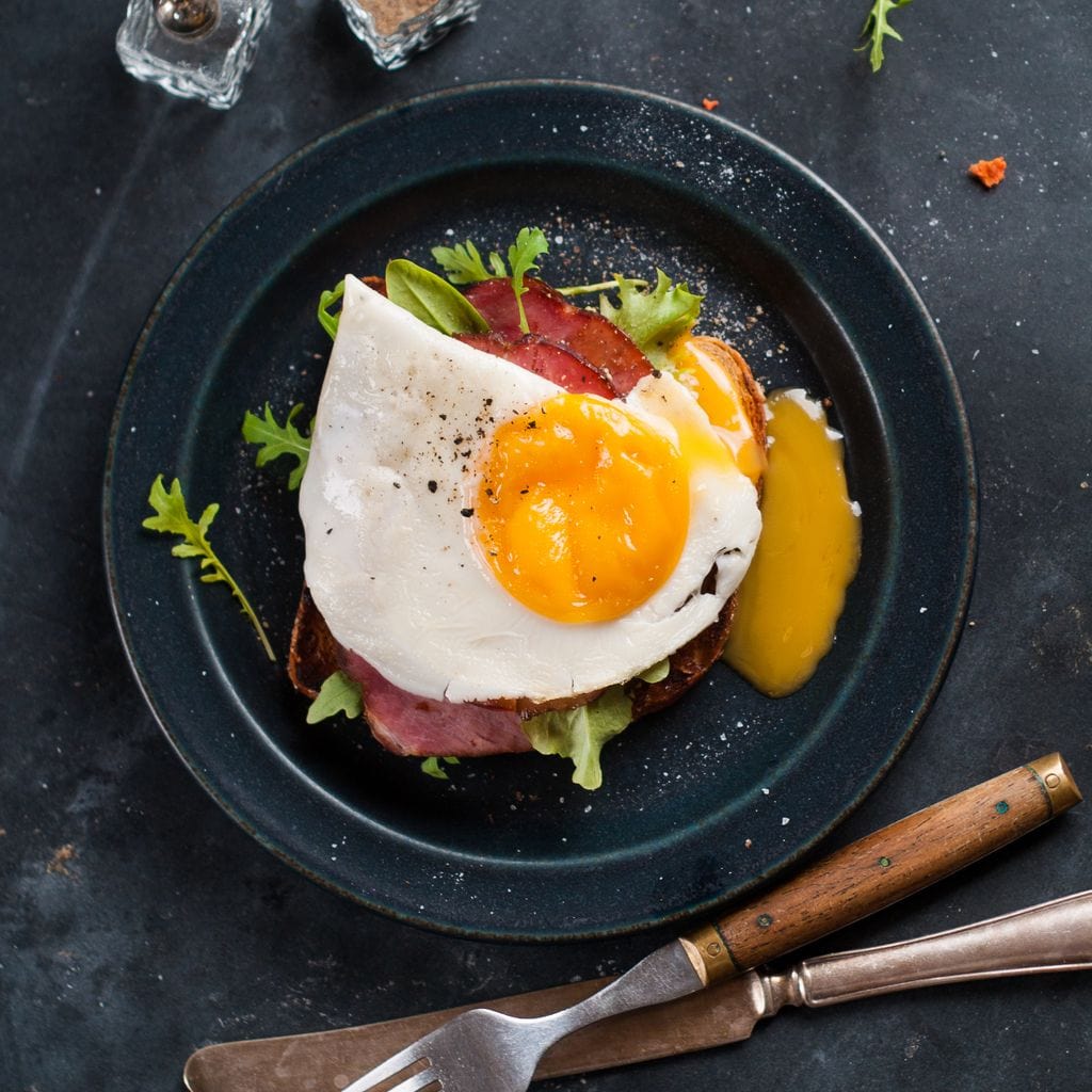 tablou canvas cu o farfurie cu un ou prajit si bacon formand un mic dejun simplu, pe o masa neagra cu tacamuri si condimente